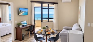 A hotel room with a bed, a desk with a laptop, a dining table with breakfast, a sofa, and a view of the sea.