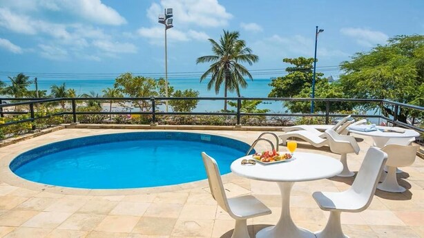 A swimming pool with lounge chairs and a table, surrounded by a railing with a view of the ocean and palm trees. - Golden Flat By Fortaleza Temporada (Fortaleza)