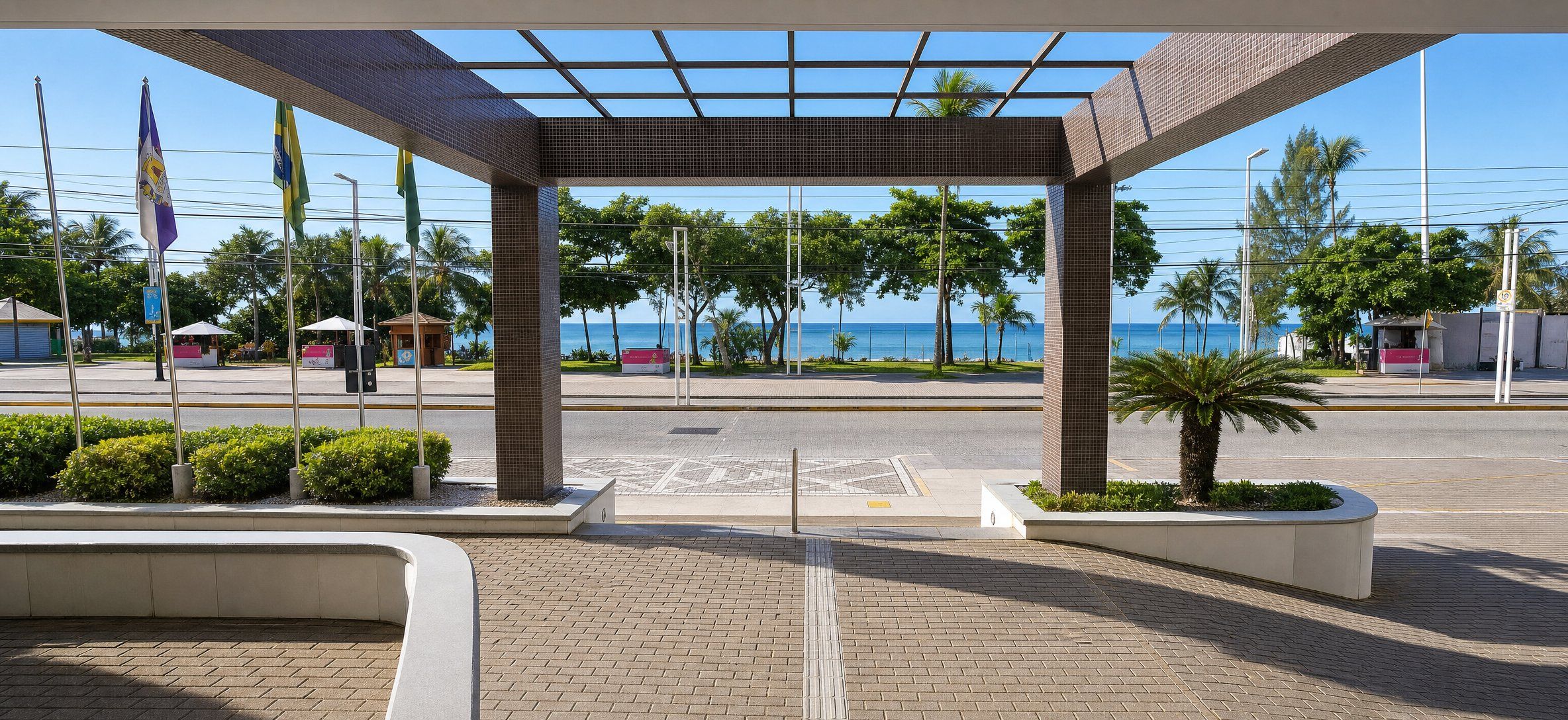 A covered pedestrian area with a view of the sea, a kiosk, and flags.