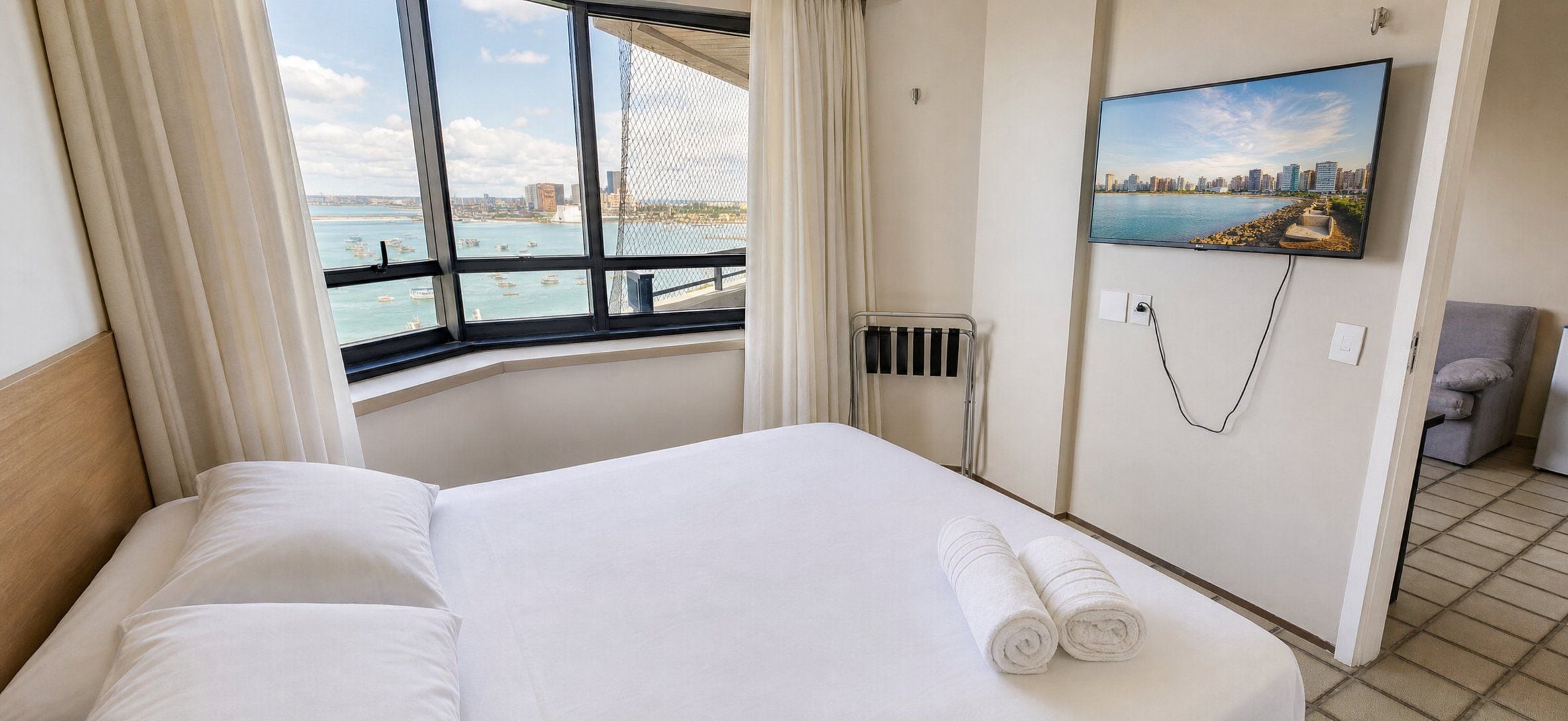 A hotel room with a large bed, a view of the sea, a painting on the wall, and a chair.