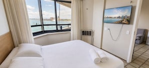 A hotel room with a large bed, a view of the sea, a painting on the wall, and a chair.