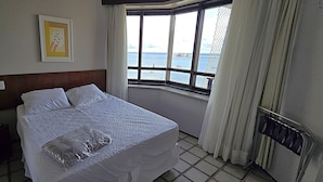 A bedroom with a bed, a nightstand, a framed picture, a window with a view of the sea, and a radiator. - Golden Flat By Fortaleza Temporada (Fortaleza)