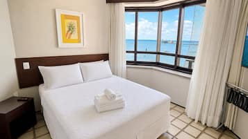 Habitación de hotel con cama, mesita de noche, control remoto, cuadro enmarcado, ventana con vista al mar y televisor montado en la pared.