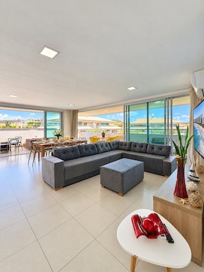 A spacious living area with a sectional sofa, a dining table with chairs, and a large window offering a view of buildings and greenery. - Cobertura no Golf Ville Deck Privativo com Jacuzzi e Churrasqueira (Aquiraz)