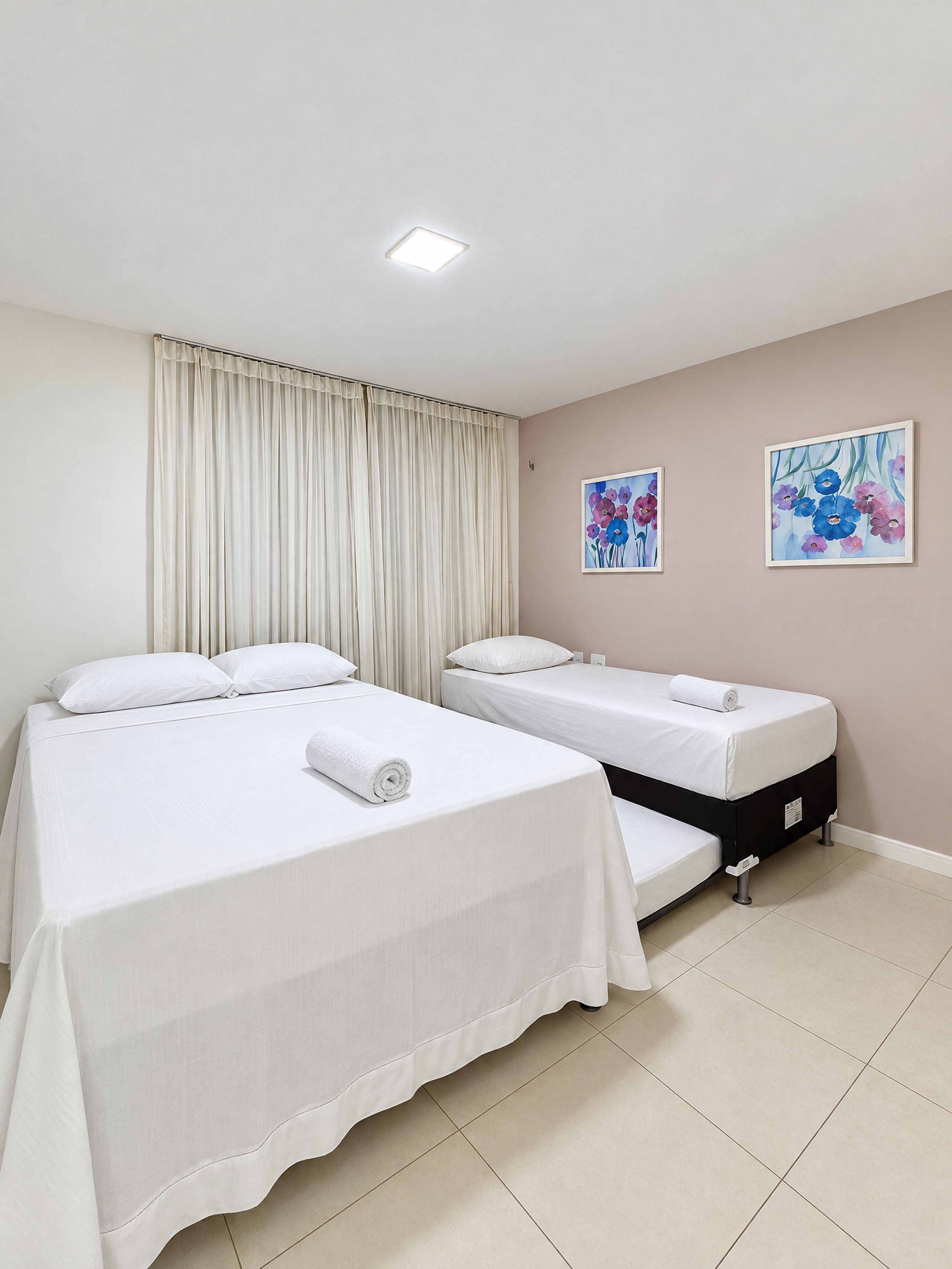 Quarto de hotel com duas camas, uma cortina e dois quadros florais na parede.