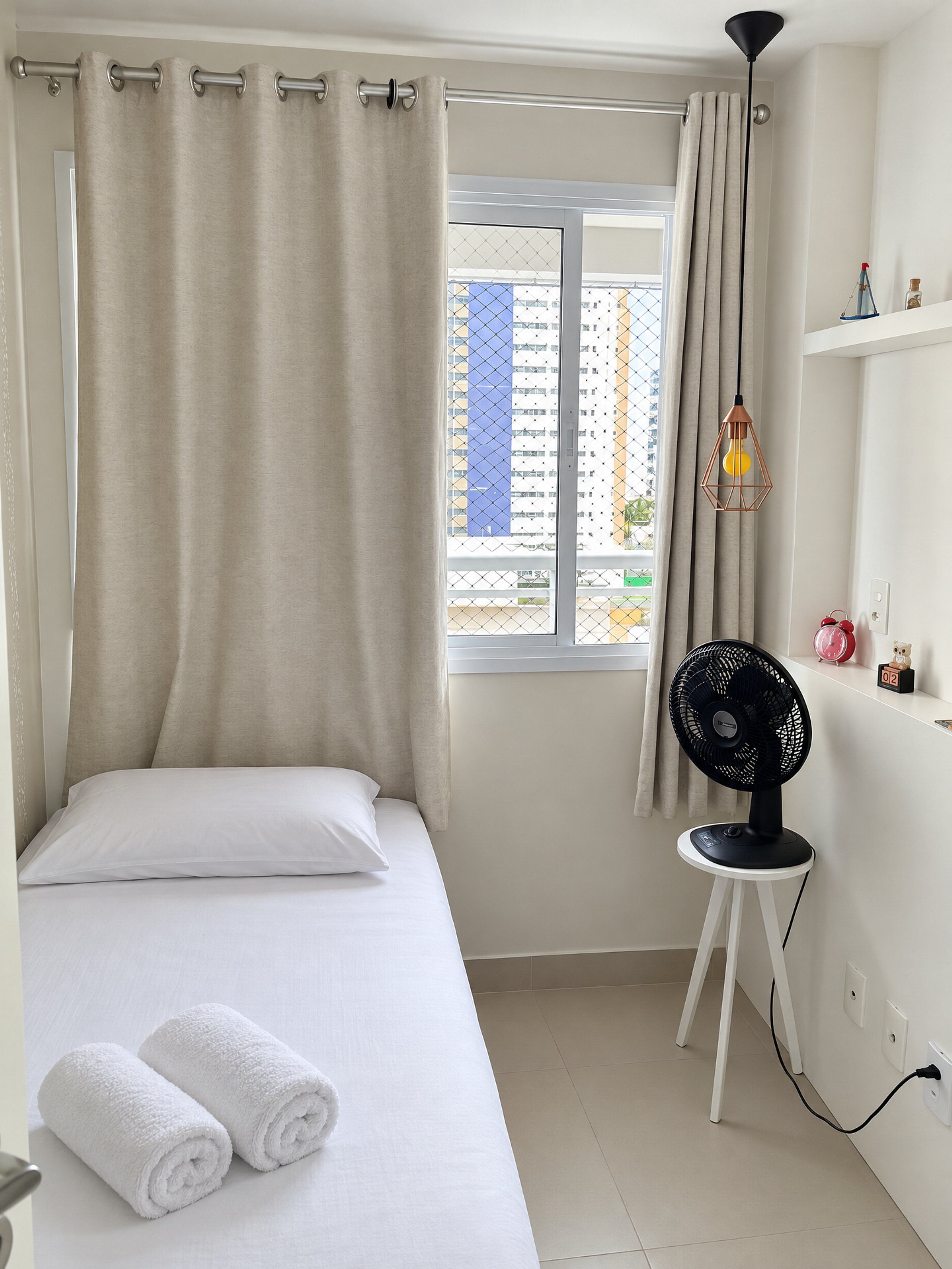A bedroom with a bed, a window with curtains, a standing fan, and a small table with a clock.