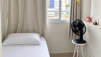 A bedroom with a bed, a window with curtains, a standing fan, and a small table with a clock.