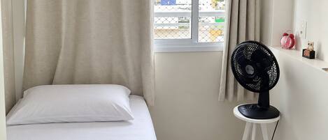 Une chambre à coucher avec un lit, une fenêtre avec des rideaux, un ventilateur sur pied et une petite table avec une horloge.
