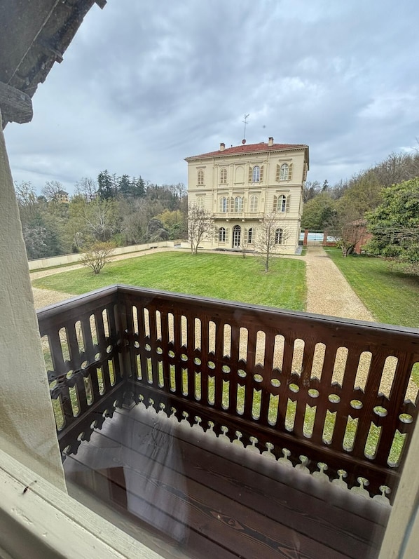 A balcony with a view of a large, classical building with a red roof, surrounded by a well-maintained lawn and trees.