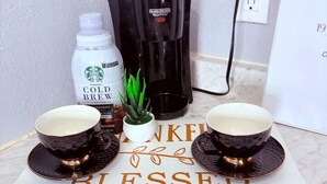 Eine Küchenarbeitsplatte mit einer Kaffeemaschine, einer Starbucks Cold Brew Flasche, einem kleinen Topfpflanzen und zwei Kaffeetassen auf einer Unterlage mit der Aufschrift 'KNIFE BLESSED'.