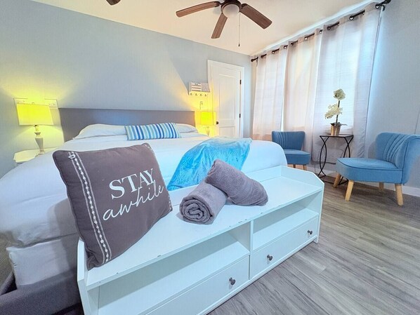 Une chambre d’hôtel avec un lit, une table de chevet, une chaise, un ventilateur de plafond et une fenêtre avec des rideaux.