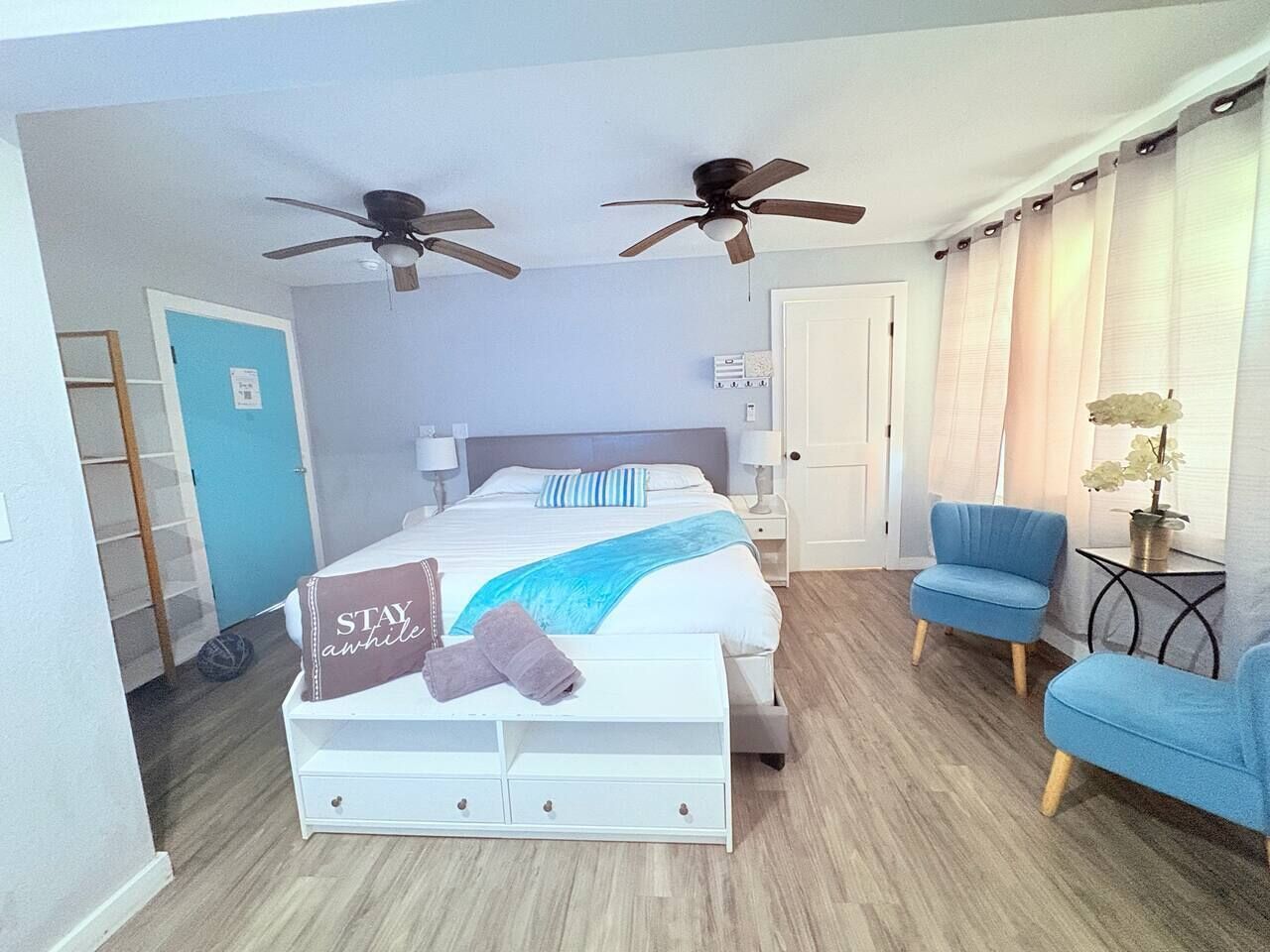 Une chambre à coucher avec un lit, deux ventilateurs de plafond, une chaise bleue, une table de chevet et une commode.