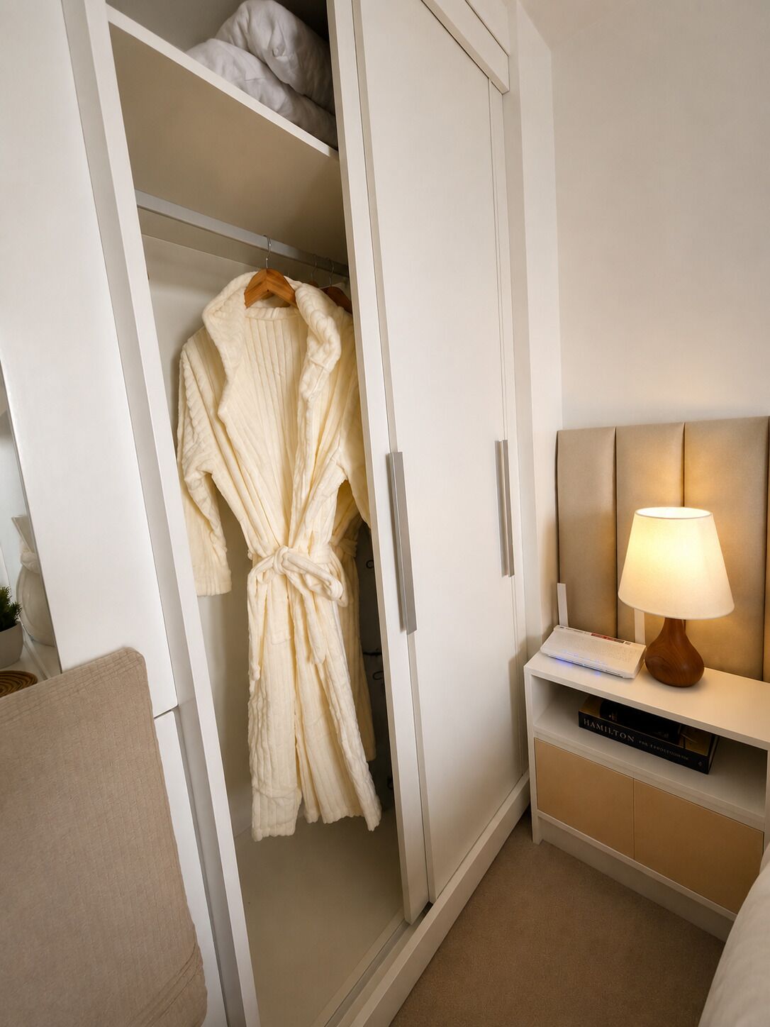 A bedroom with a white wardrobe, a bedside table with a lamp, and a book titled 'Hamilton'.