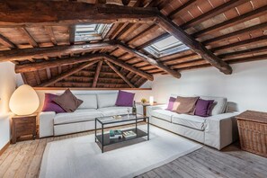 Ein Loft-Zimmer mit freiliegenden Holzbalken, einer weißen Sofagarnitur, einem Glastisch und einem Korbtisch.