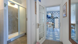 En hall som leder till ett badrum med dusch, ett köksområde med ett rutmönstrat golv och en matplats med ett bord och stolar.