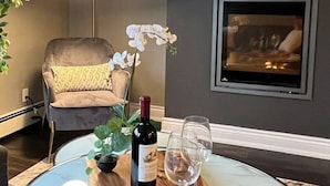 Ein runder Glastisch mit einem Holztablett, auf dem eine Weinflasche und zwei Gläser, ein Blumentopf und ein Stuhl mit Kissen in einem modernen Raum stehen.