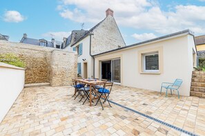 Ein Steingebäude mit einer gefliesten Terrasse, einem Holztisch mit Stühlen und einem blauen Sessel.