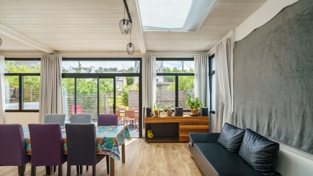 Una habitación con piso de madera, un sofá, una mesa de comedor con sillas y ventanas grandes con cortinas.