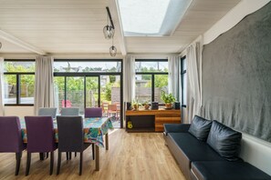 Een kamer met een houten vloer, een bank, een eettafel met stoelen en grote ramen met gordijnen.