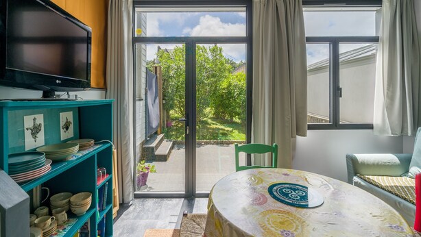 Quarto com televisão sobre uma estante azul-esverdeada, mesa com toalha de mesa florida e porta de vidro deslizante para um jardim.