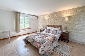 A bedroom with a stone wall, a bed with a floral duvet, bedside lamps, and a window with curtains. - Le Clos de Rouffiac - Maison pour 7 avec jardin (Carnac-Rouffiac)