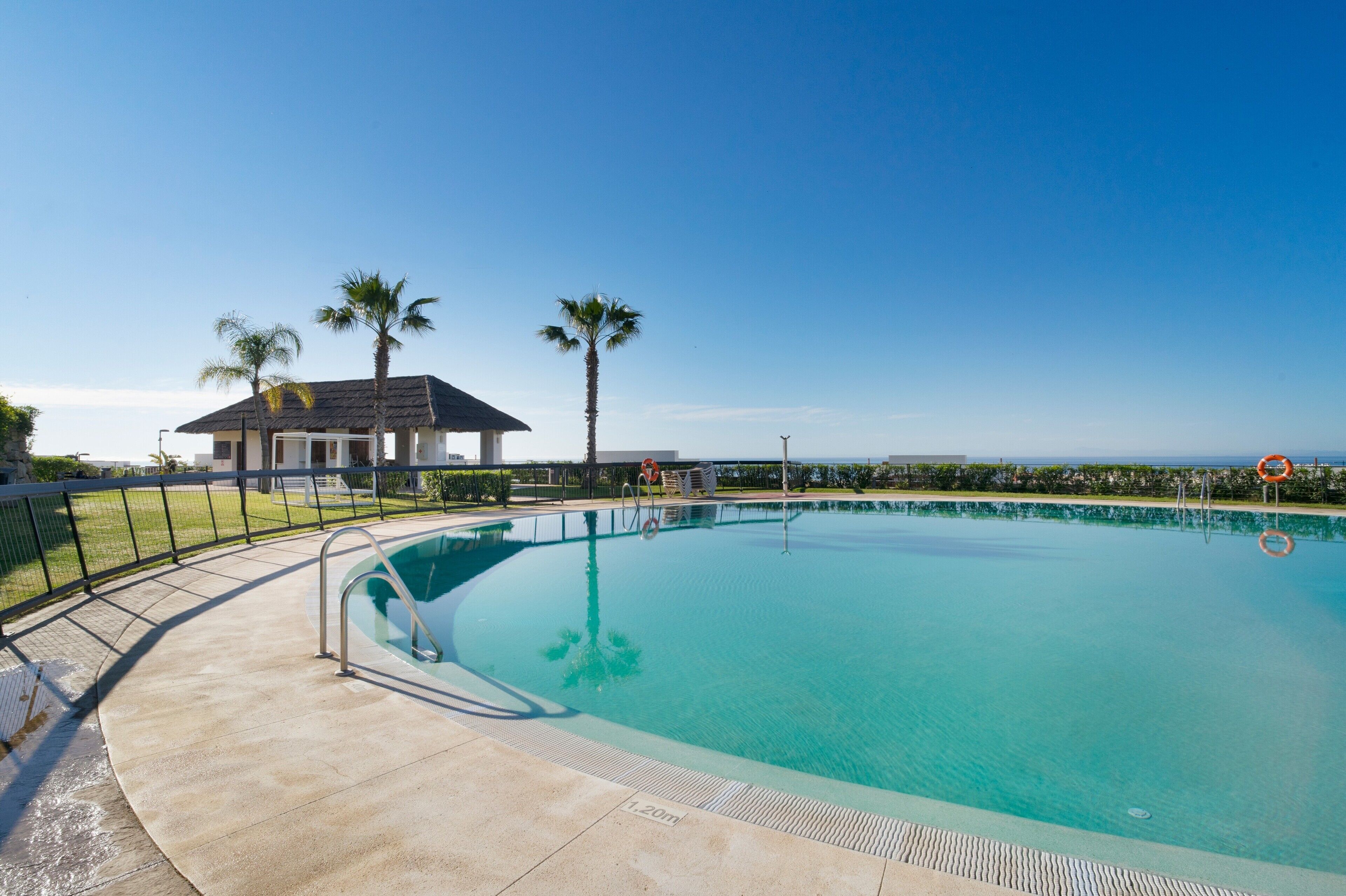 A clear blue swimming pool with a lifeguard chair, palm trees, and a building in the background.