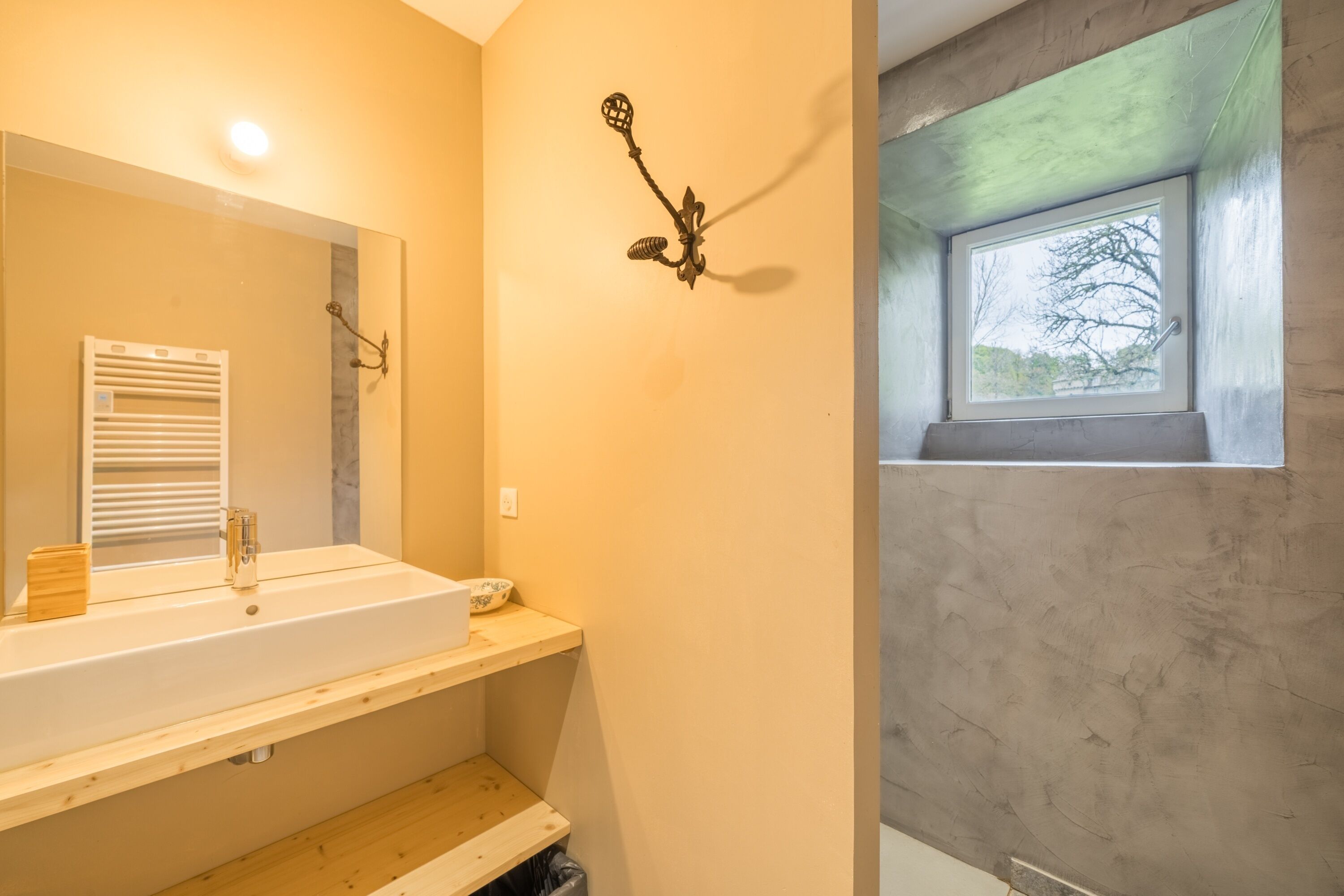 Een badkamer met een witte wastafel, houten planken, een spiegel, een wandlamp en een raam.