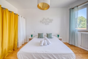Hotellihuone, jossa on suuri sänky, kaksi tyynyä, geometrisesti kuvioitu seinäkoriste ja ikkuna, jossa on verhot.