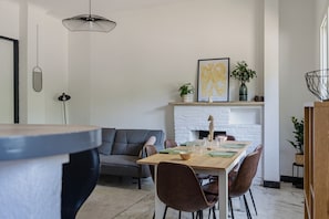 Een moderne woonkamer met een bank, een eettafel en stoelen. Er is een open haard met een witte bakstenen muur, een hanglamp en een lijst met een foto aan de wand.