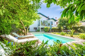 Ein Schwimmbecken mit Liegestühlen, umgeben von üppiger Vegetation und einem zweistöckigen Gebäude mit Balkon.