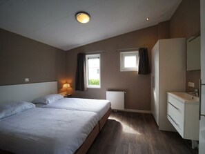 A bedroom with two beds, a window with curtains, a radiator, a bedside lamp, and a bathroom with a sink and mirror. - Holiday Home in Dordrecht With Terrace (Dordrecht)