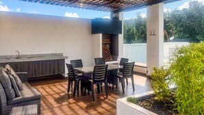 Ein überdachter Essbereich im Freien mit Tisch, Stühlen, einem Grill und Blick ins grüne Umland.