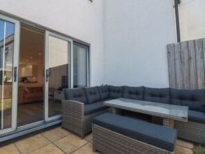 A patio with a sectional sofa, a glass door leading to a kitchen, and a wooden fence. - Cwtch Treth (Newquay)