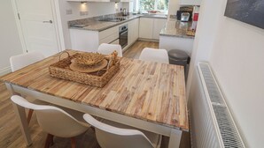 A modern kitchen with white cabinets, granite countertops, and a dining area with a wooden table and white chairs. - Cwtch Treth (Newquay)
