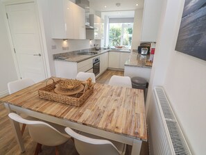 A modern kitchen with white cabinets, granite countertops, and a dining area with a wooden table and white chairs. - Cwtch Treth (Newquay)
