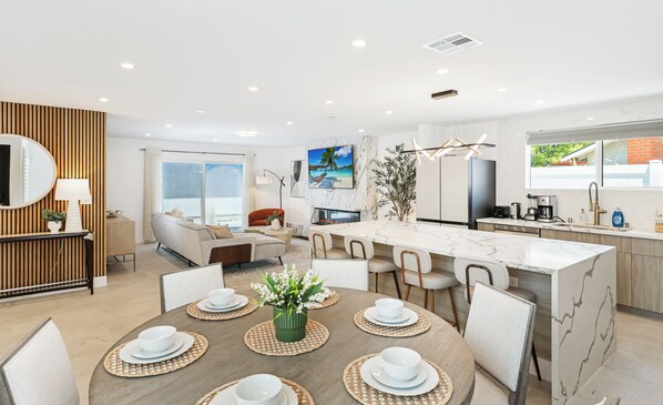 A modern kitchen with a marble island, a dining area with a round table, and a living space with a sofa and a TV.