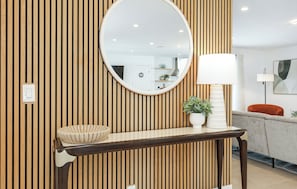 A modern bathroom with a wooden slat wall, a round mirror, a white basin, a white lamp, and a grey sofa.