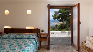 A bedroom with a bed, bedside lamps, a small table with a plant, and a view of the sea through an open door. - Domus Gallesi by Wonderful Italy (Aglientu)