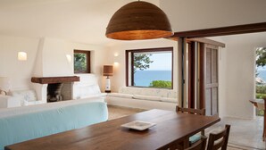 A modern living room with a wooden dining table, a sofa, a fireplace, and a large window with a view of the sea. - Domus Gallesi by Wonderful Italy (Aglientu)