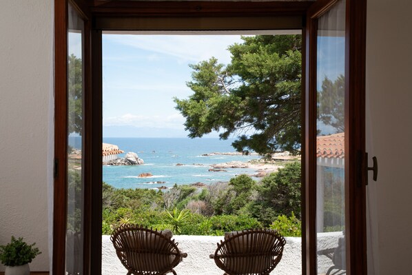 Blick von einem Balkon mit zwei Korbstühlen auf eine Küstenlandschaft mit Bäumen und dem Meer.