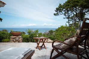 Eine Terrasse mit Blick auf das Meer, einem Holztisch, einem Stuhl und einem Steinpflanzgefäß.