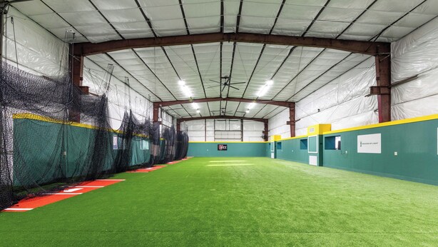 Un complexe sportif intérieur doté d’un terrain synthétique, de zones délimitées par des filets et d’un tableau d’affichage.