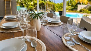 Mesa de jantar posta para seis pessoas, com pratos brancos, copos transparentes e talheres, com vista para uma piscina e palmeiras.
