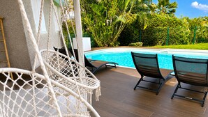 Uma área à beira da piscina com cadeiras de macramé e espreguiçadeiras, um deck de madeira e uma piscina.