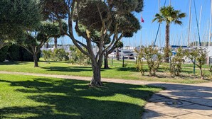 A park with a tree-lined path, a grassy area, and a marina with boats in the background. - Sol-y-Days Jardins du port large terrace and swimming pool (Le Grau-du-Roi)