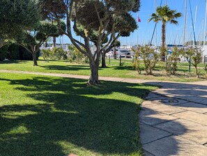 Un parc avec un sentier bordé d’arbres, une pelouse et un port de plaisance où l’on aperçoit des bateaux au loin.