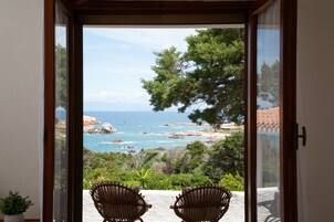 Vista desde una habitación con dos sillones de mimbre orientados a un paisaje costero con árboles y el mar.