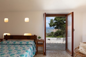 Ein Schlafzimmer mit Bett, Nachttischlampen, einem kleinen Tisch mit Pflanze und Blick aufs Meer durch eine offene Tür.