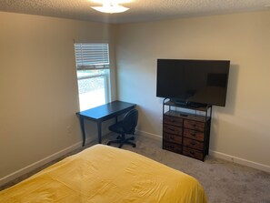 Ein Schlafzimmer mit Bett, Schreibtisch, Stuhl, Fernseher auf einem Schrank und einem Fenster.