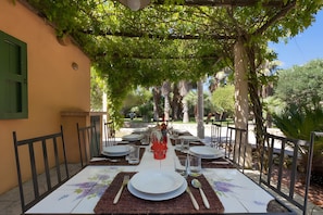 Un'area pranzo all'aperto con una pergola, tavoli apparecchiati con piatti, posate e bicchieri, circondata dal verde.